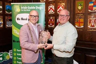 IGD CEO Tim O'Mahony presenting a glass vase to Lord Mayor McAdam in front of an Irish Guide Dogs for the Blind banner, with coats of arms on the wall behind.