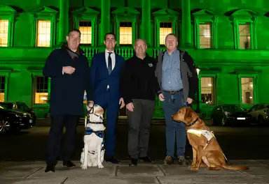 Four men stand in a line with two service dogs in front of a brightly lit green building at night.