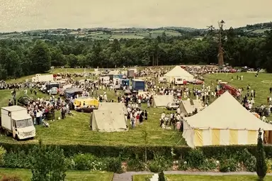 A lively outdoor festival with tents, vehicles, and crowds of people on a grassy field surrounded by trees and hills in the background.