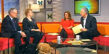 Four people sit on a TV talk show set with a guide dog resting on the floor. A screen displays "Ireland AM" in the background.
