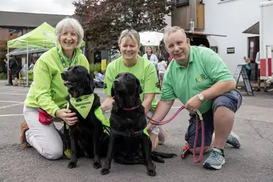 Marie Ryan (Limerick Branch) and Ger and Liam Hennessy (Kinsale Branch) in green IGD shirts are kneeling with two black Labradors wearing "Ambassador Dog" vests
