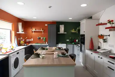 Modern kitchen with orange and green walls, a wooden island, appliances, and tableware set for a meal. Bright and organized space.