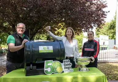 CEO Tim O'Mahony, with Maeve O'Connell TD and Cork Camogie's Amy O'Connor smiling beside a raffle drum at an Irish Guide Dogs event, celebrating their 50th anniversary with a trophy on a green table.