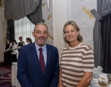 Minister Jerry Buttimer in a blue suit and Tara Mullally (daughter of Jim Dennehy) in a striped shirt smile for the camera in an elegant room with people and a table with cups in the background.