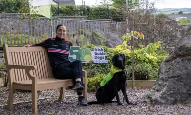 Amy sits on a bench holding a "50 Irish Guide Dogs 1976-2026" sign, with a black ambassador dog wearing a yellow vest in a garden setting.