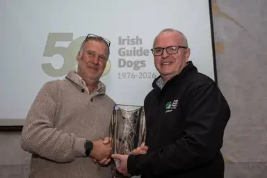 Peter and IGD CEO Tim O'Mahony shake hands, holding a glass award, standing in front of an Irish Guide Dogs 50th anniversary banner.