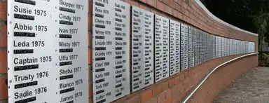 A brick memorial wall displays white plaques with names and years in chronological order from 1975 onwards, each with Braille translations beneath the text.