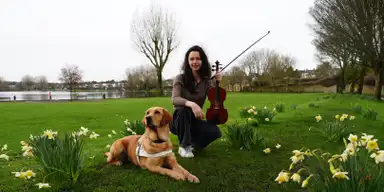 Mairead Hickey kneels in a park, holding a violin, with a guide dog lying beside her among blooming daffodils. A lake and trees are in the background.