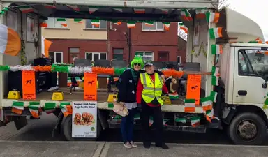 Two volunteers in festive attire stand in front of an Irish-decorated parade float promoting Irish Guide Dogs for the Blind, adorned with green, white, and orange flags and banners