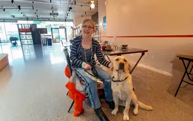 A woman wearing glasses and a striped blazer sits on a stool next to a yellow Labrador Retriever in a modern, well-lit indoor space.