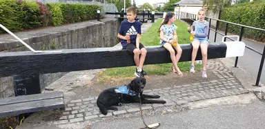 Three children sit on a black beam by a canal, enjoying drinks. Poncho, with his Assistance Dog jacket on, lies on the cobblestone path below.