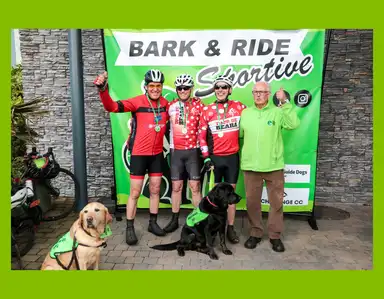 Cyclists in red outfits pose with two guide dogs and a man in green, standing in front of a "Bark & Ride Sportive" sign.