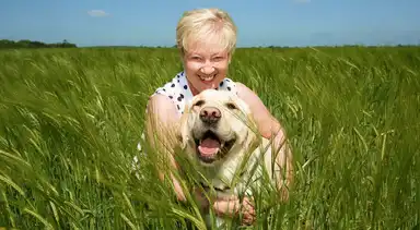 A smiling woman with short blonde hair wearing a polka-dot blouse poses with a yellow Labrador Retriever in a green grain field under a blue sky.