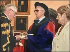 An academic ceremony where Jim is in a graduation gown receiving a diploma from an official, with a woman standing nearby. Portraits hang on the wall.