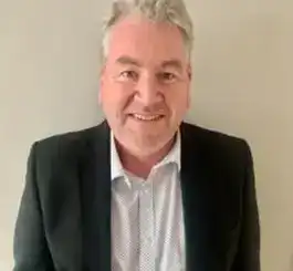 A middle-aged man with gray hair smiling at the camera while wearing a dark blazer over a light blue button-up shirt.