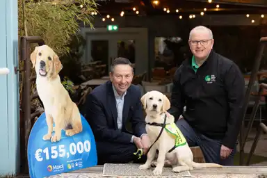 The image shows two men standing with two dogs, one a golden retriever and the other a service dog, in front of a building with festive lights, and a large sign indicating a donati