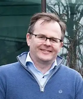 Pat in glasses wearing a blue zip-up jacket over a collared shirt, standing outdoors with trees and glass building in the background.