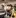 Two service dogs, a yellow Labrador and a brown Labrador, sit side-by-side on black tires in a sandy training area, both wearing white harnesses and collars.