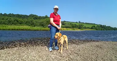 Jo in a red polo shirt and white cap stands on a pebbly beach with a yellow Labrador Retriever on a leash, with a calm lake and forested hillside in the background under clear