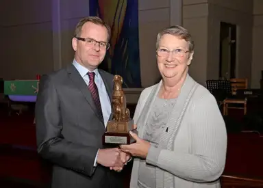 Two people shake hands while holding a bronze award statue in what appears to be a formal church or institutional setting.