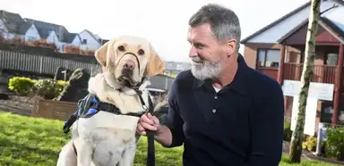 A middle-aged man with gray hair and beard smiles while holding the leash of a yellow Labrador Retriever wearing a blue service vest in a residential backyard setting.