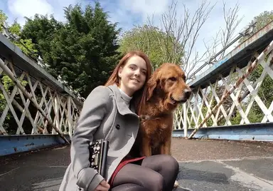 A smiling young woman with reddish-brown hair crouches beside a golden-brown dog on a bridge, holding a guitar while posing for the camera.