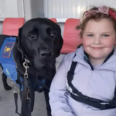 A young girl with flowers in her hair smiles next to a black Labrador service dog wearing a blue vest, seated together on red chairs.