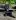 A person wearing sunglasses and dark clothing sits on a wooden bench while a black dog lies on the ground in front of them.