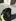 A black Labrador Retriever wearing a bright yellow service vest sits on a paved patio area, looking up at the camera.