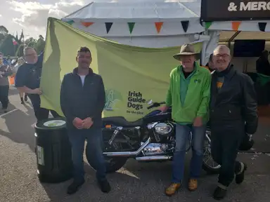 Three men stand in front of a motorcycle and a green banner for Irish Guide Dogs, outdoors at an event.