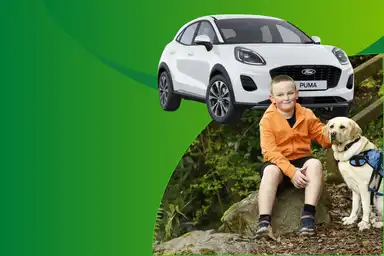 A white Ford Puma car on a green background above a boy in an orange jacket sitting on rocks with a guide dog.