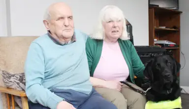Ann and her husband sitting together indoors with a black guide dog, Finlay, wearing a yellow harness by their side.