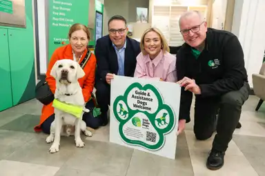 The image shows a group of people, including a guide dog and staff members from the Irish Guide Dogs, standing together and holding a sign that welcomes guide and assistance dogs.