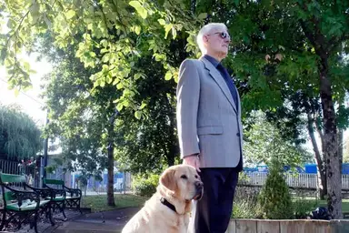 Jim in sunglasses stands with a yellow Labrador, under trees in the grounds of the Training Centre. Benches and greenery surround them.