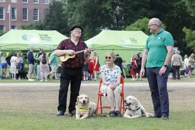 Gus Dorrington playing ukulele, Paula Dorrington sitting with guide dogs, Tim O'Mahaony CEO standing, in Merrion Square with green tents in the background.