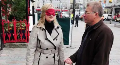A woman in a trench coat, blindfolded with a red cloth, is guided by a man in a brown jacket on a city street.