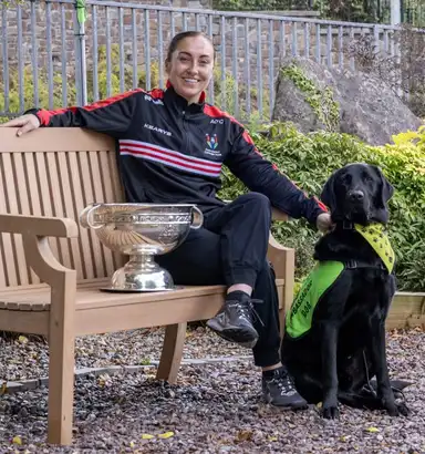 Amy in sportswear sitting on a bench with a silver trophy, smiling, and a black labrador wearing a green vest beside them.