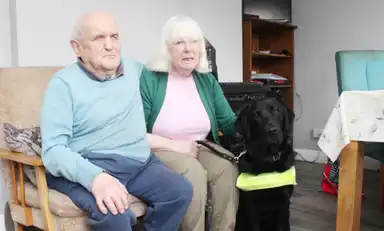 Ann and her husband Eamon sit on a sofa with guide dog Finlay wearing a neon harness, in a cozy living room with a table and bookshelf.