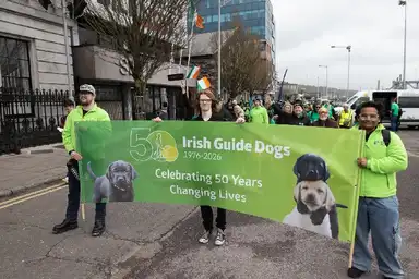 Group holding a "Irish Guide Dogs: Celebrating 50 Years" banner, marching in a parade with flags and people in green.