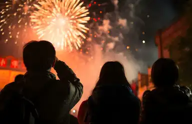 Silhouetted figures watch a vibrant fireworks display illuminate the night sky in warm orange and gold tones.