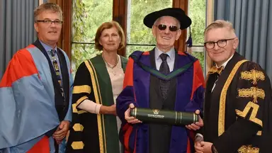 Four people including Jim Dennehy in academic robes pose indoors; one holds a diploma, wearing sunglasses and a graduation cap. A window and greenery are in the background.