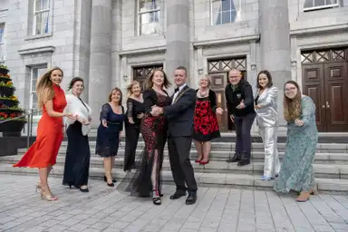 A group of people in formal attire pose on steps in front of a grand building, smiling and gesturing towards the camera.