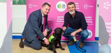 Two men in business casual attire kneel beside a black Labrador service dog wearing a yellow vest at a Dublin Airport Parking promotional event.