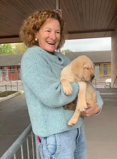 Sonia smiling while holding a sleeping yellow Labrador puppy, wearing a blue sweater and jeans, standing outdoors under a wooden roof.