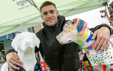 IGD Ambassador Jack Crowley in a black jacket poses with two dog sculptures, one white and one colorful, under a green tent.