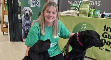 Geraldine in a green shirt kneels beside two black dogs with guide dog vests at a booth for guide dog awareness.
