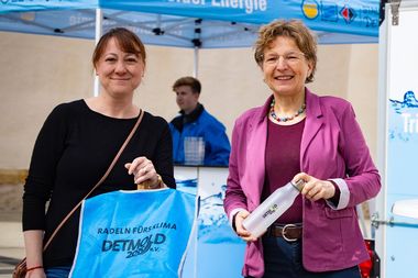 Zwei Frauen lächeln, während sie eine blaue Warnweste und Wasserflaschen am Stand von Detmold Energie halten. Im Hintergrund sind Banner und ein Mitarbeiter sichtbar.