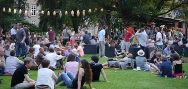 Menschen sitzen im Park, im Hintergrund HÜtten und Lichterketten.