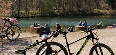 Fahrräder stehen im Vordergrund, dahinter Menschen an einem Fluss.