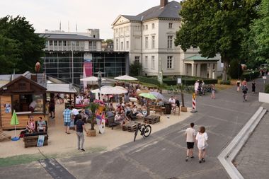 Menschen sitzen an einer Strandbar mitten in der Stadt am Landesmuseum.